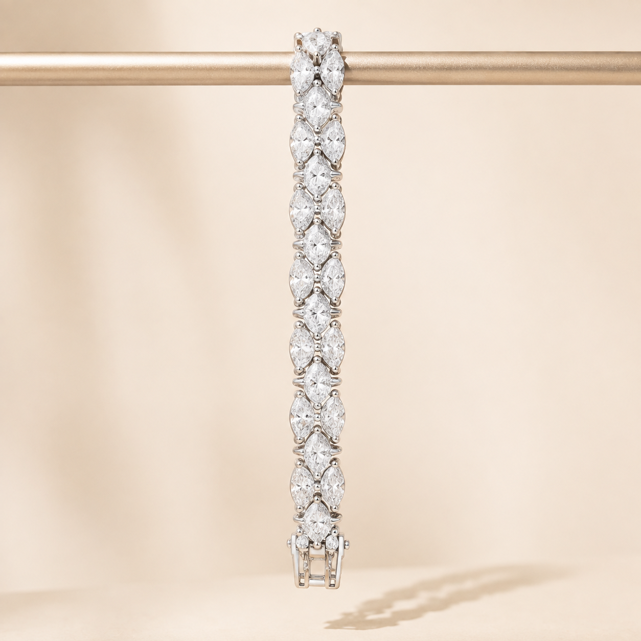 A diamond tennis bracelet with marquise-cut stones set in a silver-toned metal, hanging vertically from a gold display rod against a warm beige background.