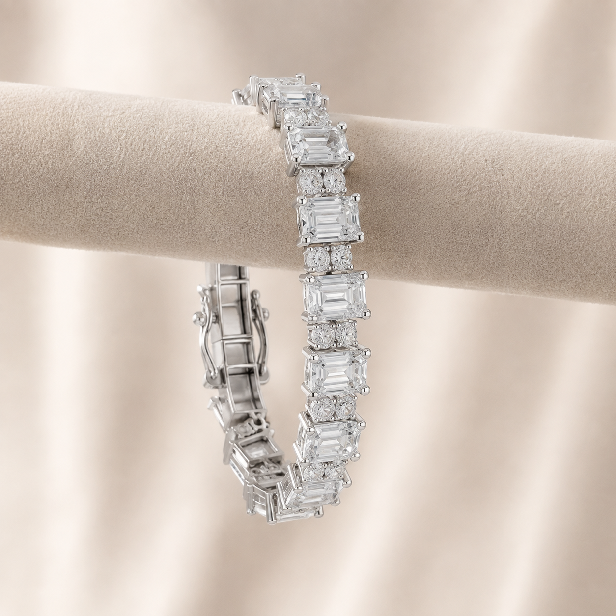 A silver-toned tennis bracelet with emerald-cut clear stones, displayed on a beige fabric jewelry stand against a soft beige background.