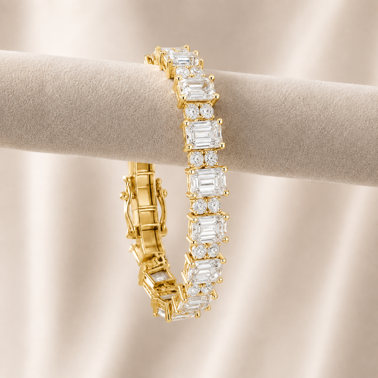 A gold tennis bracelet with baguette-cut clear stones, displayed on a beige jewelry stand against a soft beige fabric background.