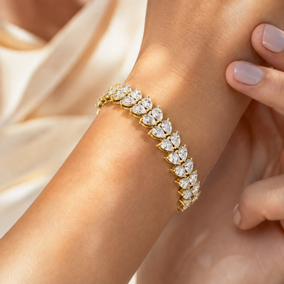 A close-up of a gold tennis bracelet with marquise-cut clear stones worn on a woman's wrist, with soft cream satin fabric in the background.