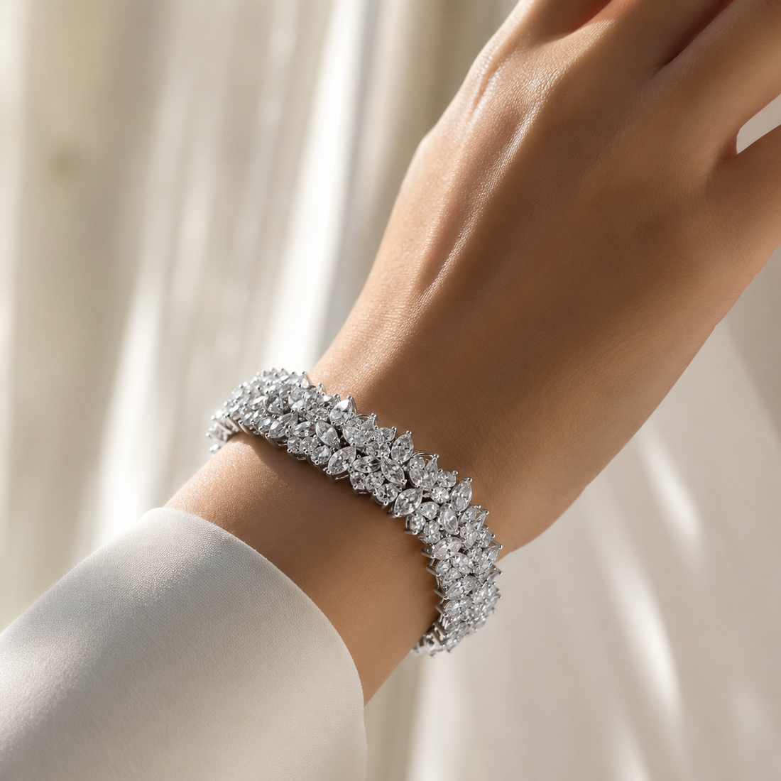 A close-up of a person's wrist wearing a sparkling silver-toned bracelet adorned with clear gemstones, set against a soft, light cream-colored curtain background.