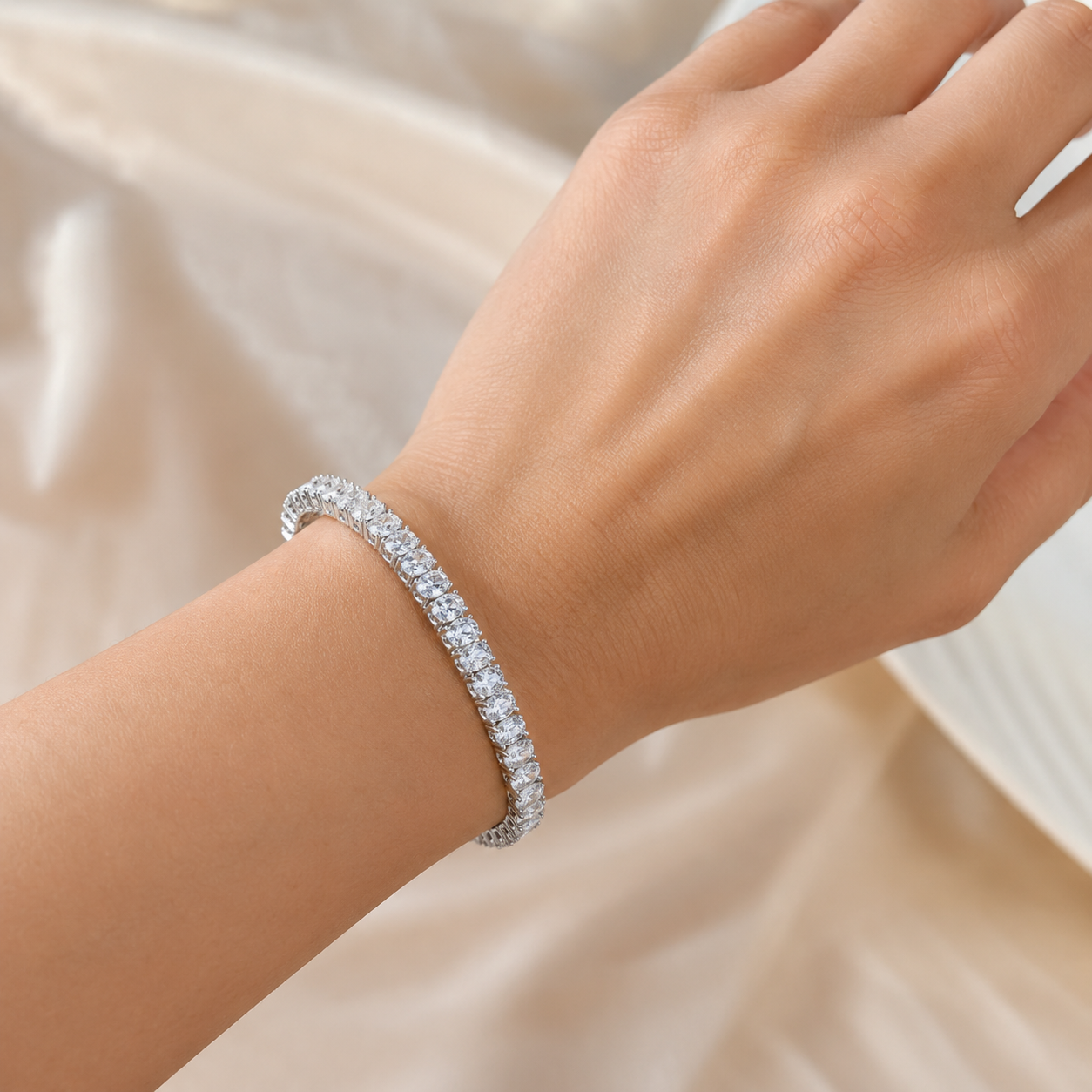A close-up of a person's wrist wearing a sparkling silver tennis bracelet with clear stones, set against a soft beige satin fabric background.