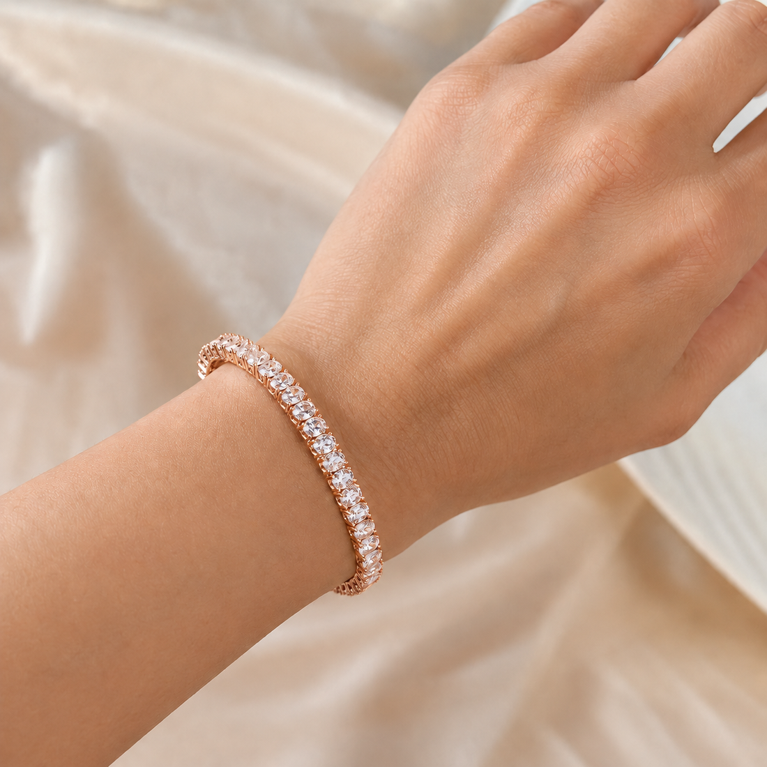 A close-up of a person's wrist wearing a rose gold tennis bracelet with clear round-cut stones, set against a soft beige satin fabric background.