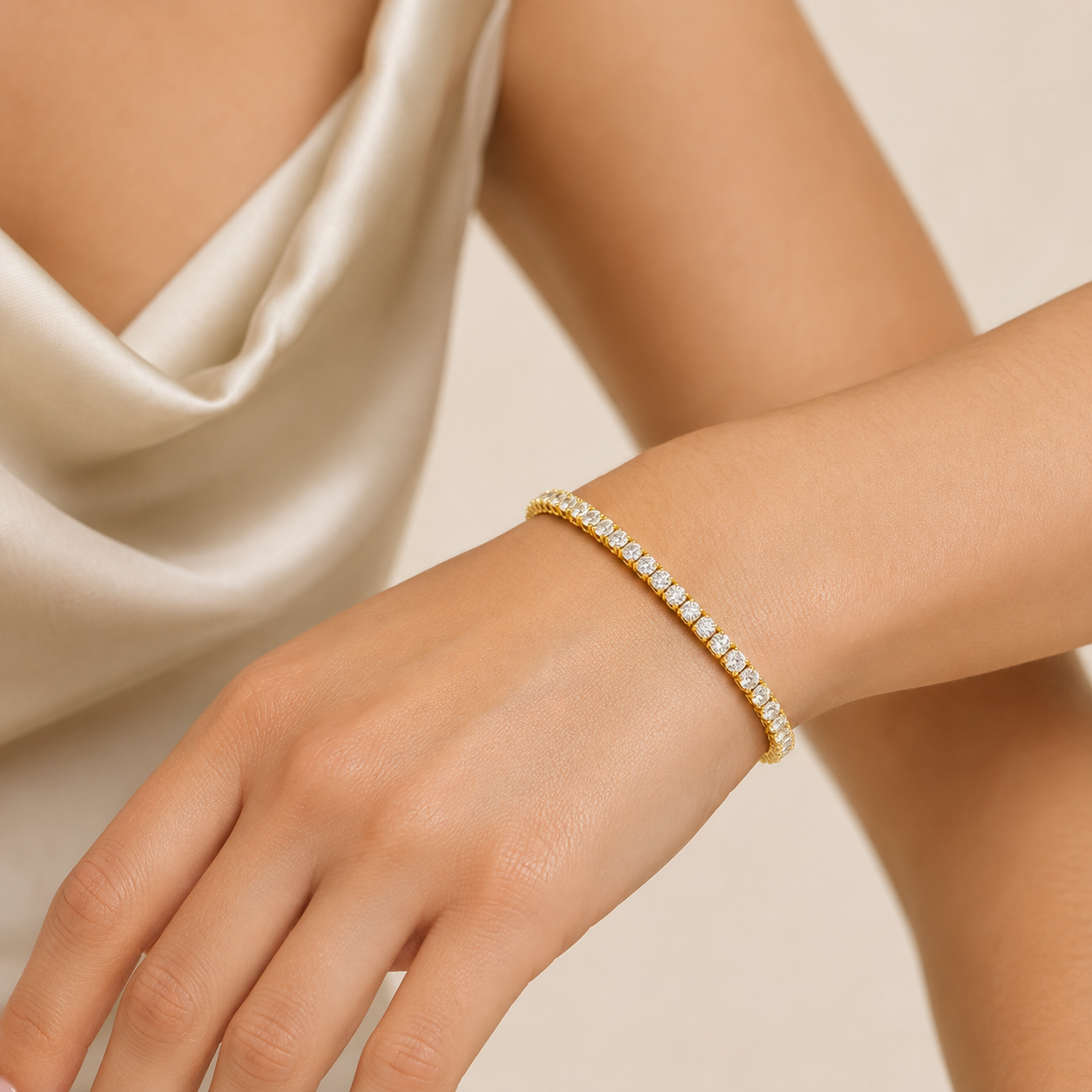 A delicate gold tennis bracelet with small clear stones, worn on a woman's wrist. The image features a close-up of the hand and wrist, with a soft beige satin dress in the background.