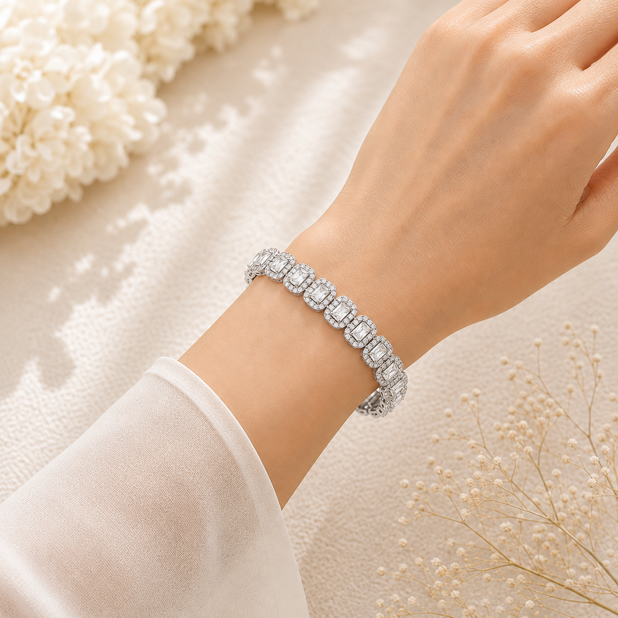 A close-up of a woman's wrist wearing a silver-toned tennis bracelet with rectangular clear stones, styled with soft white flowers on a light textured fabric background.