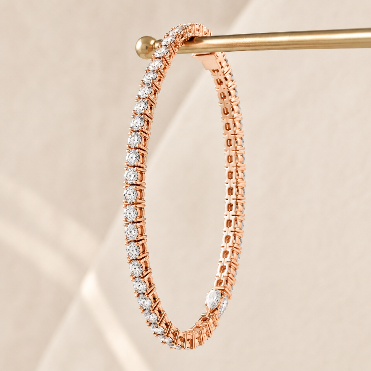 A rose gold tennis bracelet with a continuous line of round-cut clear stones, hanging from a gold display stand against a warm beige background.