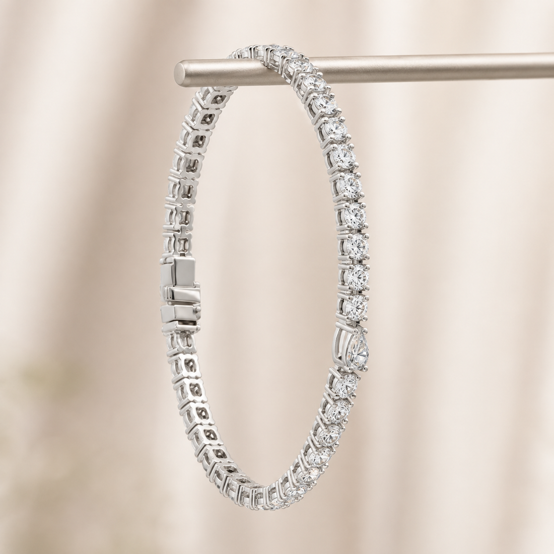 A silver tennis bracelet with clear round-cut gemstones, hanging from a silver display bar against a soft beige fabric background.