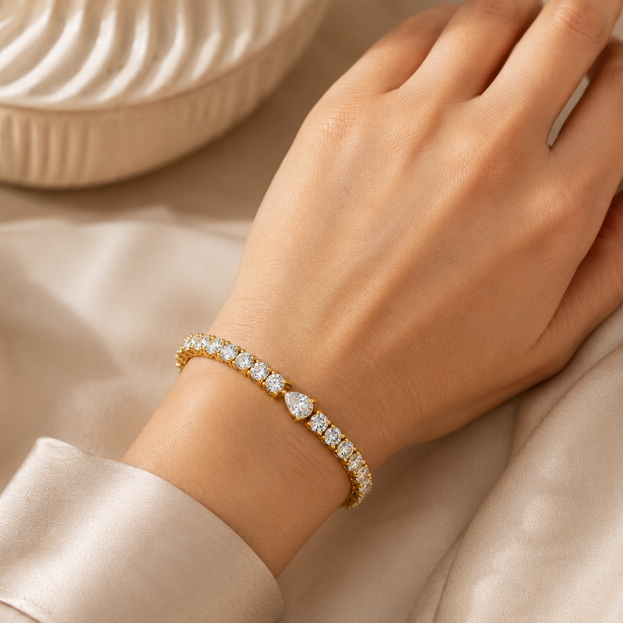 A close-up of a person's hand wearing a delicate gold tennis bracelet adorned with sparkling clear gemstones, set against a soft beige fabric background.