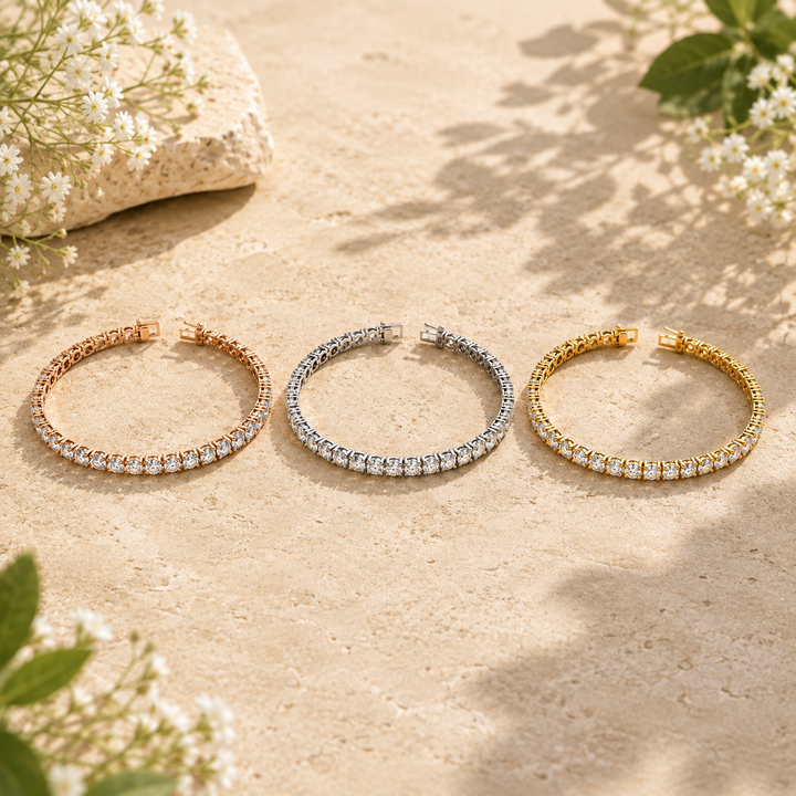 Three tennis-style cuff bracelets with pavé-set clear stones are arranged on a sandy surface outdoors. The bracelets are shown in rose gold, silver, and yellow gold tones.