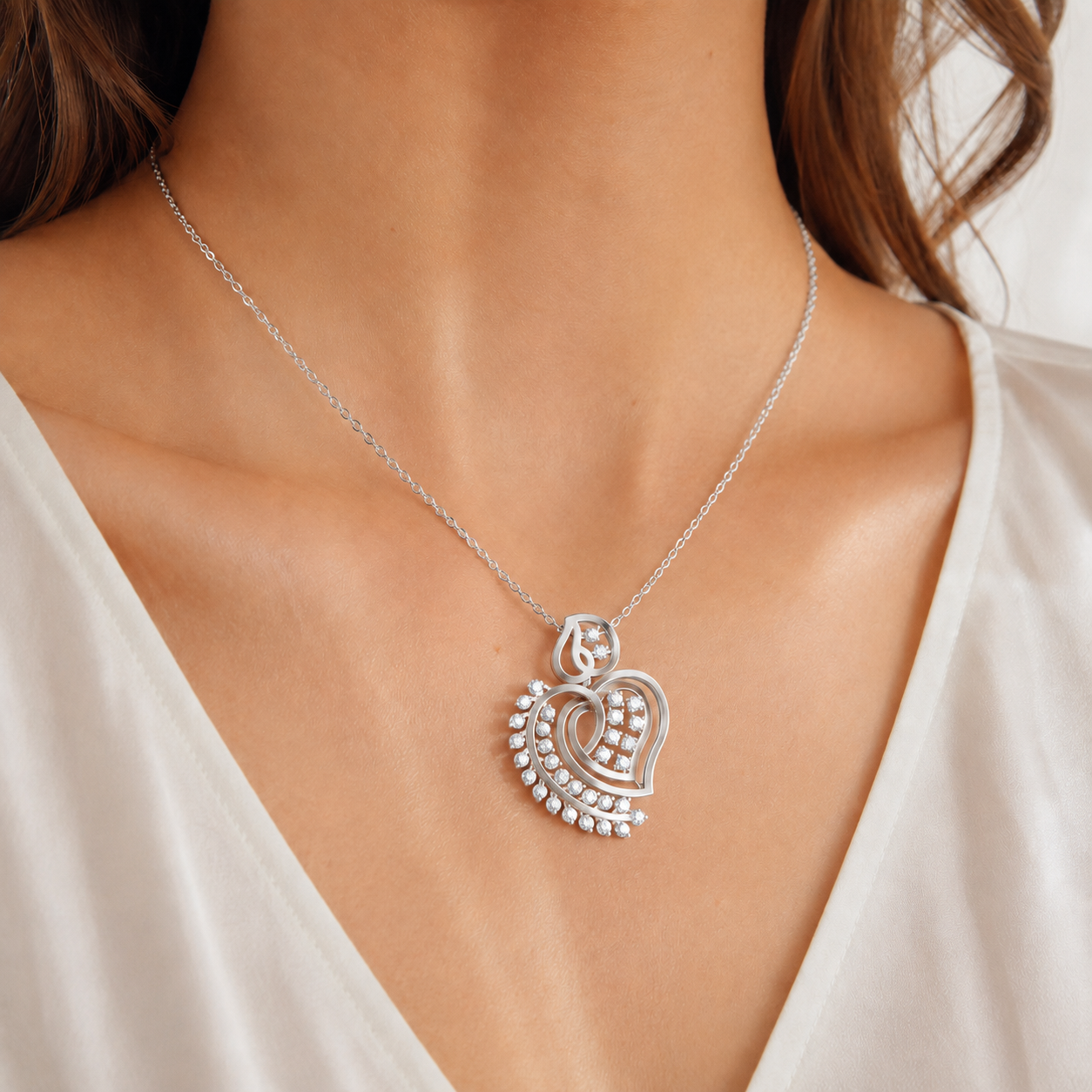A close-up of a woman's neck and chest wearing a delicate silver chain necklace with a heart-shaped pendant. The pendant features a central heart motif surrounded by small clear stones, set against a soft white top.