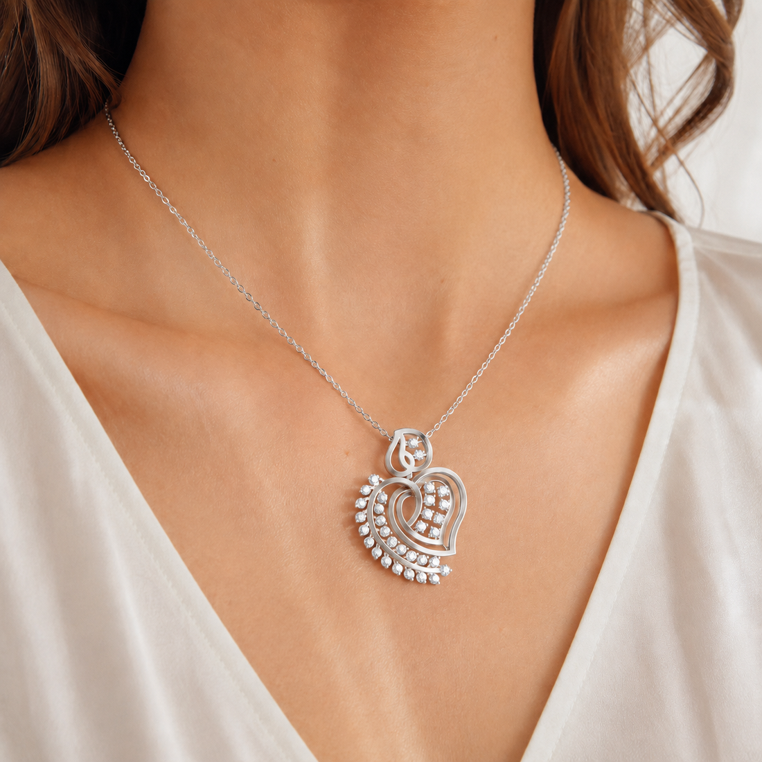 A close-up of a woman's neck and chest wearing a delicate silver chain necklace with a heart-shaped pendant. The pendant features a central heart motif surrounded by small clear stones, set against a soft white top.