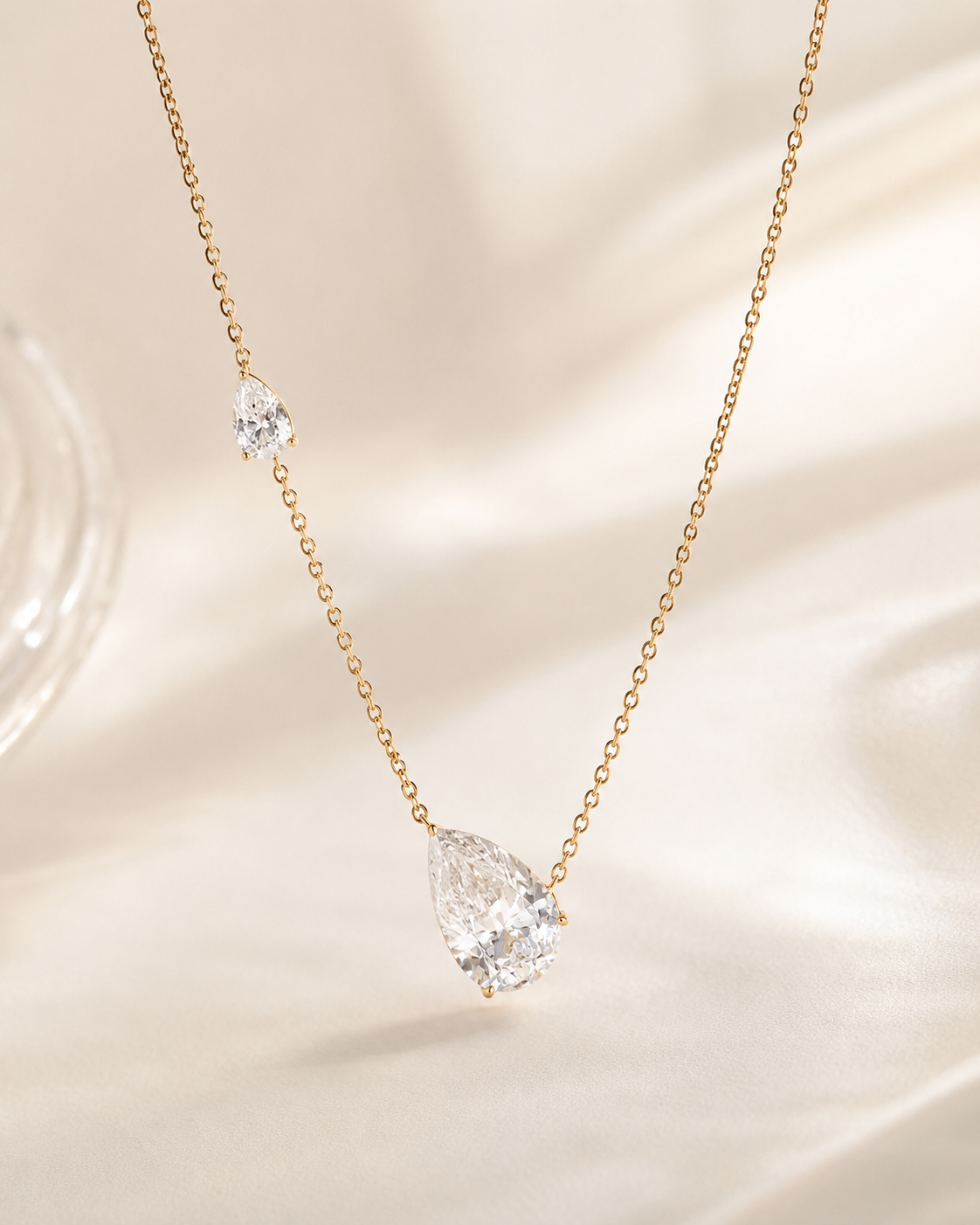 A delicate gold chain necklace featuring a large, faceted pear-cut clear gemstone pendant and a smaller matching clear gemstone accent on the opposite side, displayed on a soft beige satin fabric background.