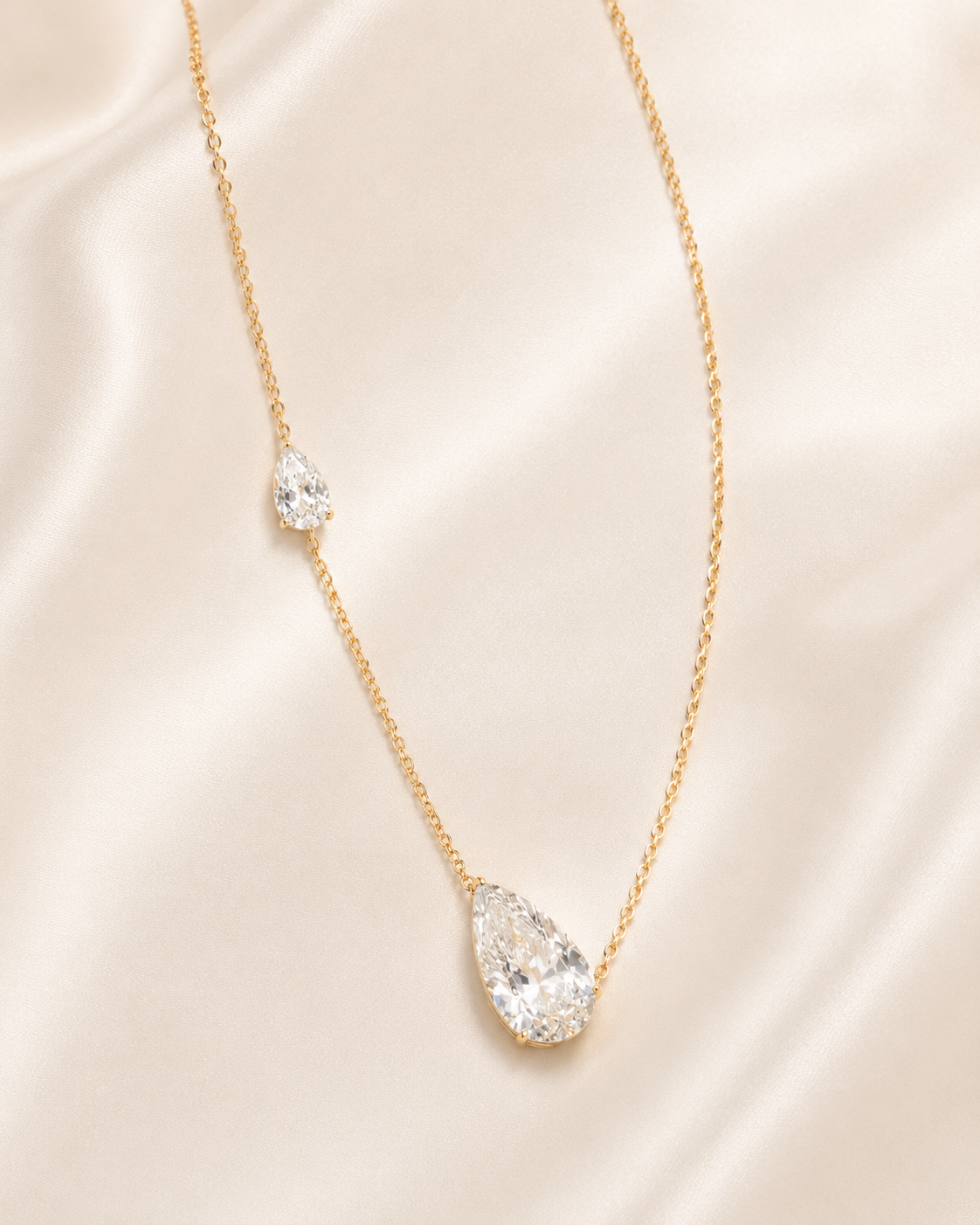A delicate gold chain necklace featuring a large teardrop-shaped clear gemstone pendant and a smaller matching teardrop accent on the chain, displayed on a soft beige satin fabric background.