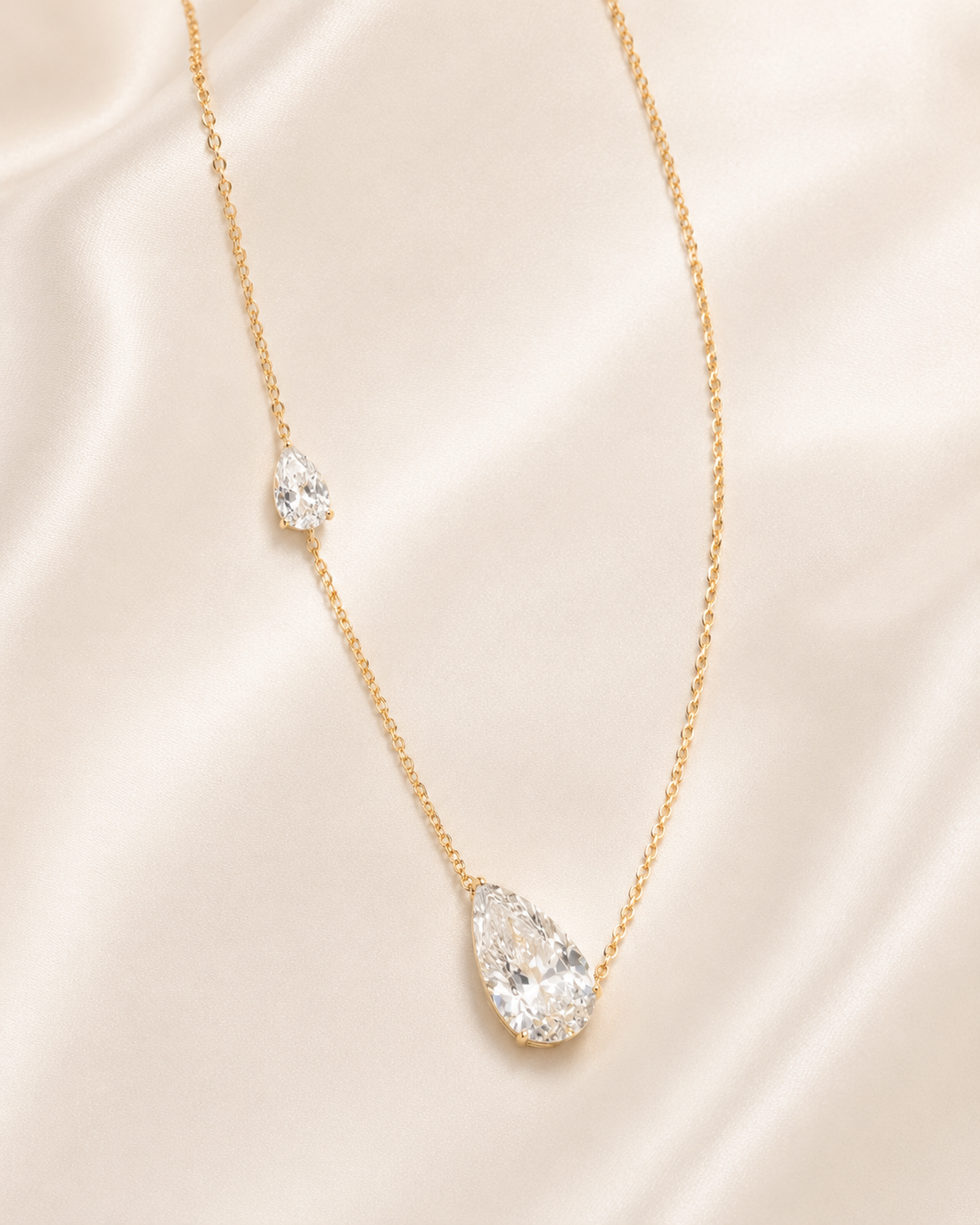 A delicate gold chain necklace featuring a large teardrop-shaped clear gemstone pendant and a smaller matching teardrop accent on the chain, displayed on a soft beige satin fabric background.