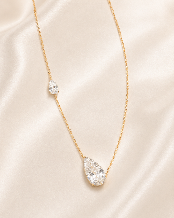A delicate gold chain necklace featuring a large teardrop-shaped clear gemstone pendant and a smaller matching teardrop accent on the chain, displayed on a soft beige satin fabric background.
