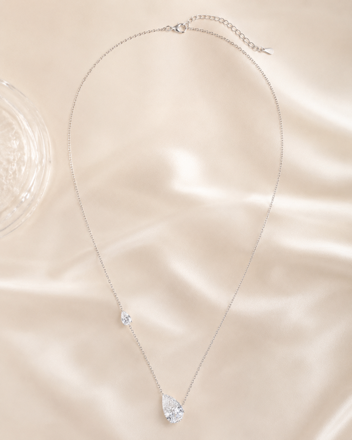 A delicate silver chain necklace with a teardrop-shaped clear crystal pendant and a small clear crystal accent, displayed on a beige satin fabric background.