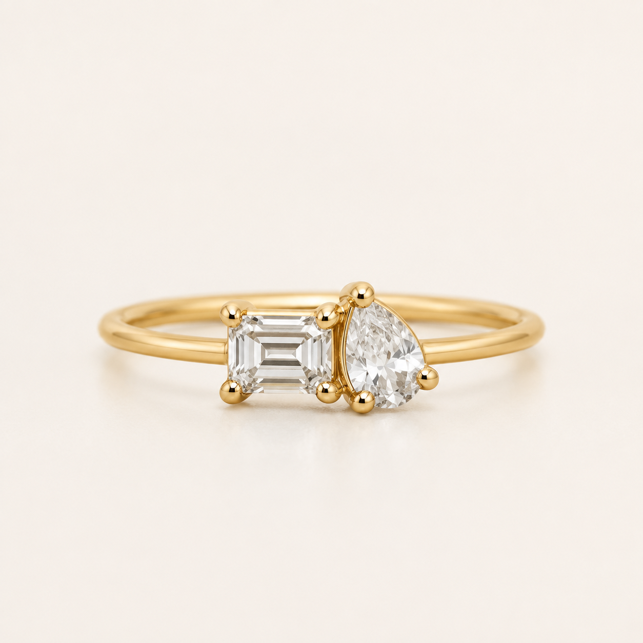 A yellow gold ring featuring an emerald-cut clear diamond on the left and a pear-cut clear diamond on the right, set on a thin gold band against a light cream background.