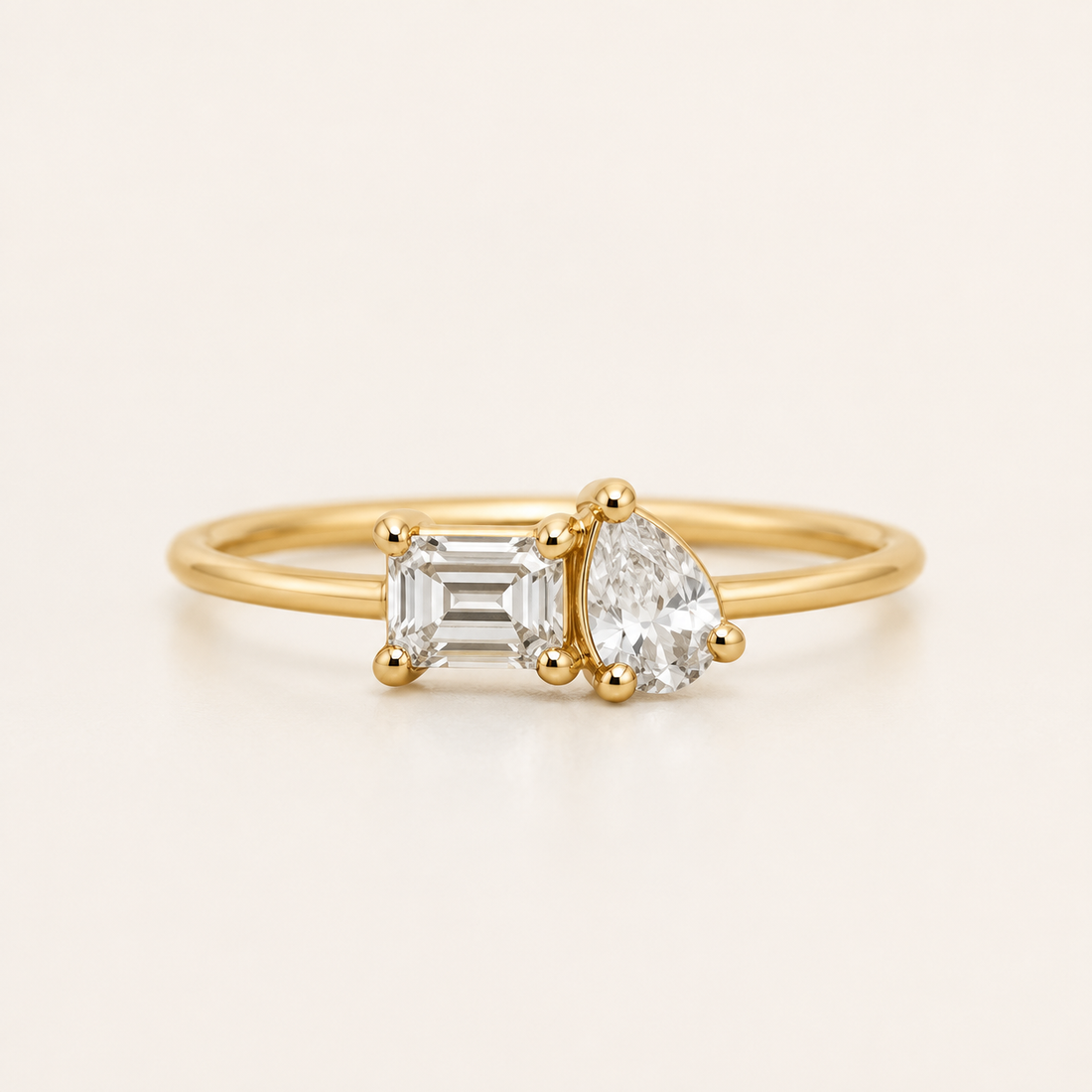 A yellow gold ring featuring an emerald-cut clear diamond on the left and a pear-cut clear diamond on the right, set on a thin gold band against a light cream background.