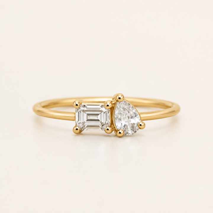 A yellow gold ring featuring an emerald-cut clear diamond on the left and a pear-cut clear diamond on the right, set on a thin gold band against a light cream background.