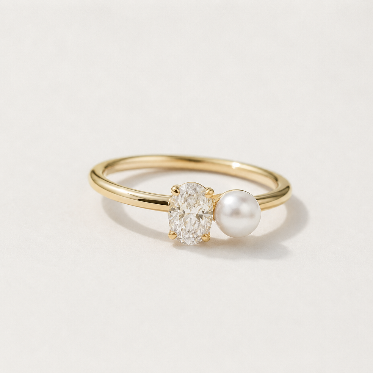 A delicate yellow gold ring featuring an oval-cut clear gemstone and a white pearl set side by side on a slim gold band, photographed on a clean white background.