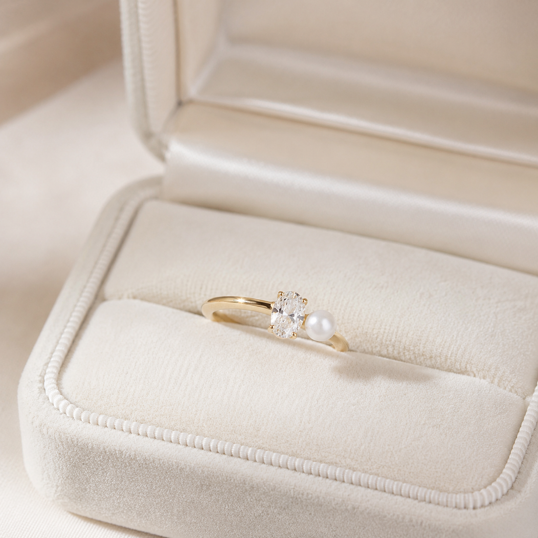 A gold ring with a single clear gemstone and a white pearl, displayed in an open cream-colored velvet jewelry box.