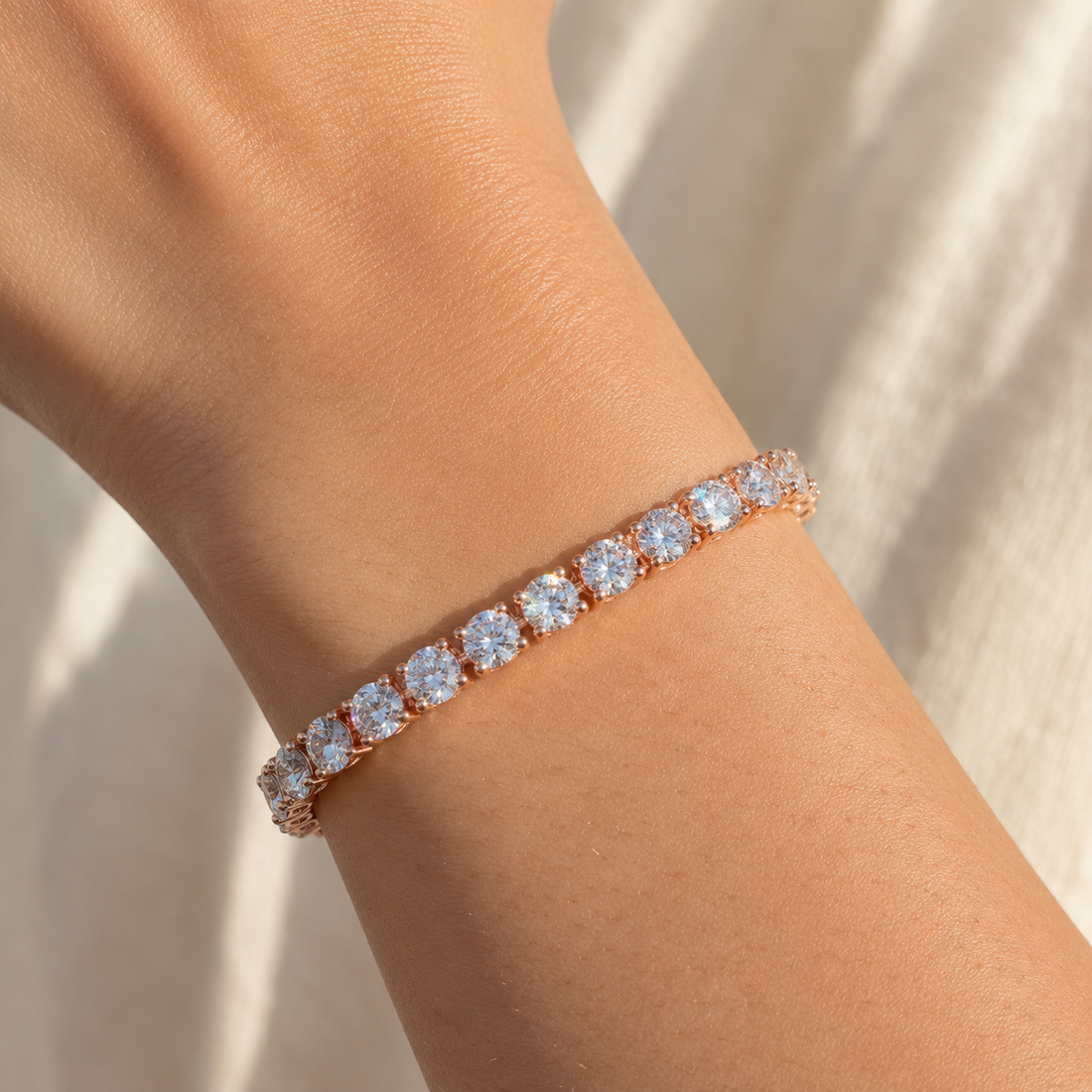 A close-up of a rose gold tennis bracelet with clear round-cut gemstones worn on a person's wrist against a soft beige fabric background.