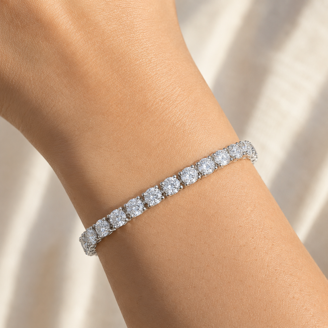A close-up of a diamond tennis bracelet on a person's wrist, set in a silver-toned metal with a warm champagne background.