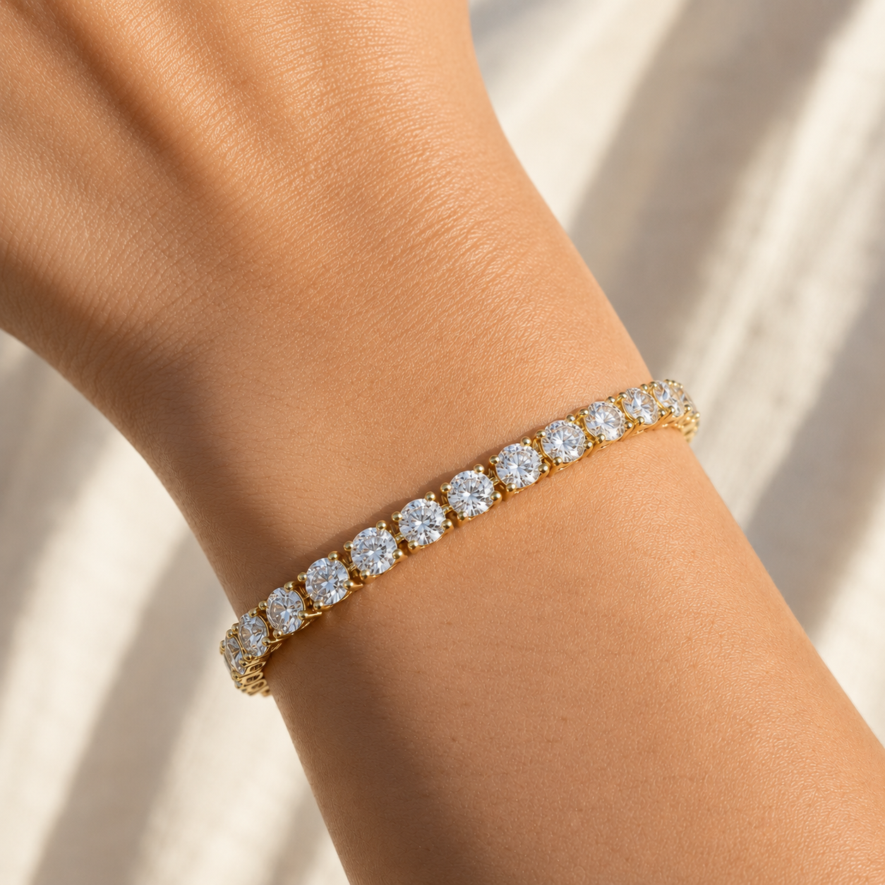 A gold tennis bracelet with round clear stones worn on a person's wrist, photographed against a softly lit light beige background.