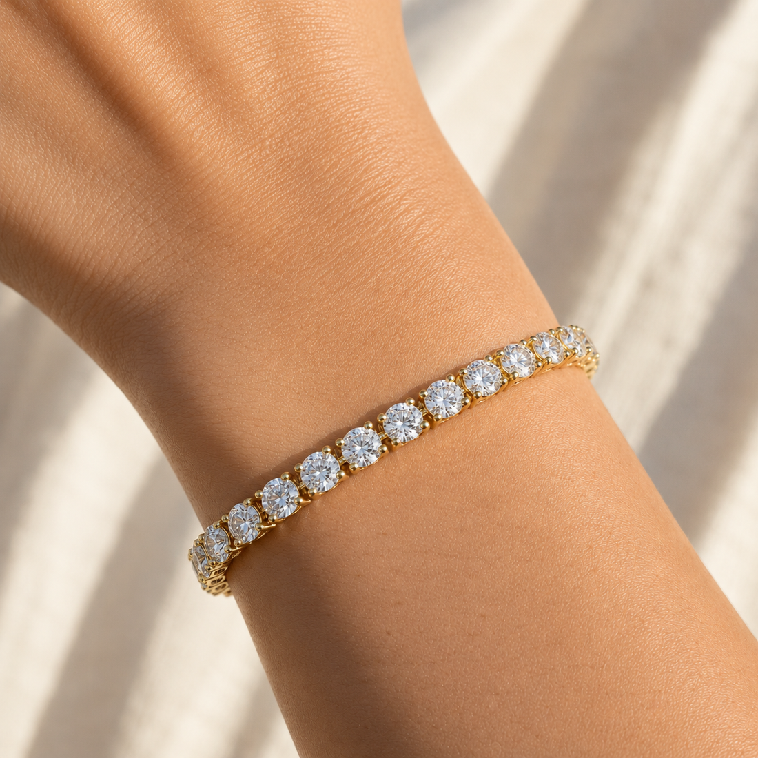 A gold tennis bracelet with round clear stones worn on a person's wrist, photographed against a softly lit light beige background.