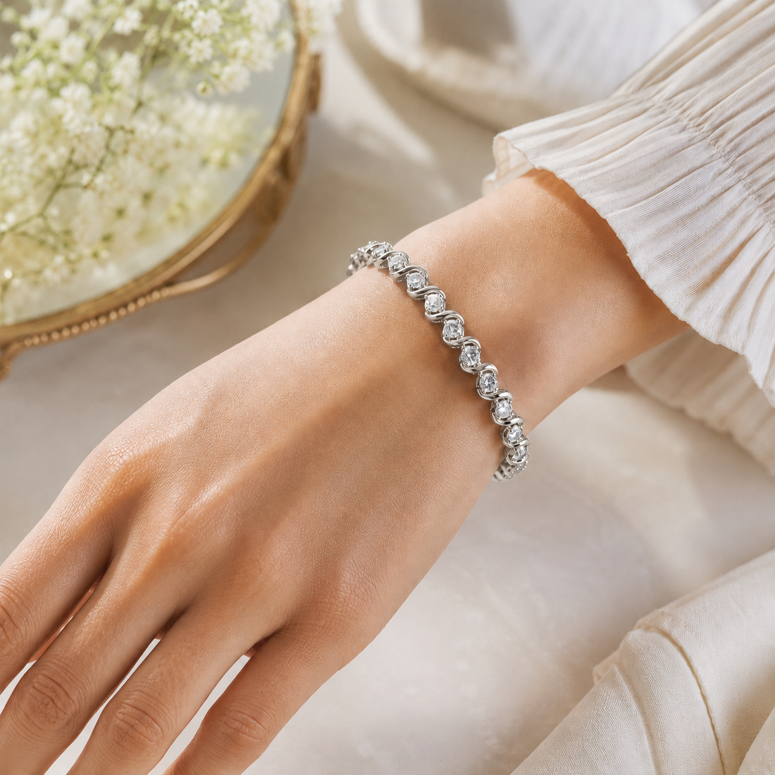 A silver-toned beaded bracelet with clear faceted stones worn on a woman's wrist, shown against a soft cream fabric background with a decorative gold-framed floral mirror partially visible.