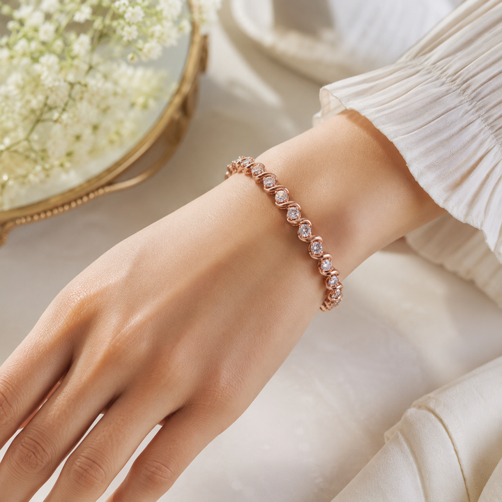 A close-up of a person's wrist wearing a delicate rose gold bracelet with clear faceted gemstones, styled with a white pleated sleeve and a soft cream background.