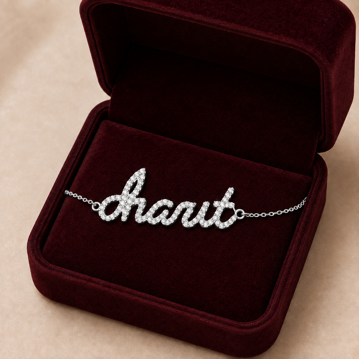 A silver script nameplate bracelet paved with small clear stones spelling charit displayed inside an open dark burgundy velvet jewelry box on a beige background.