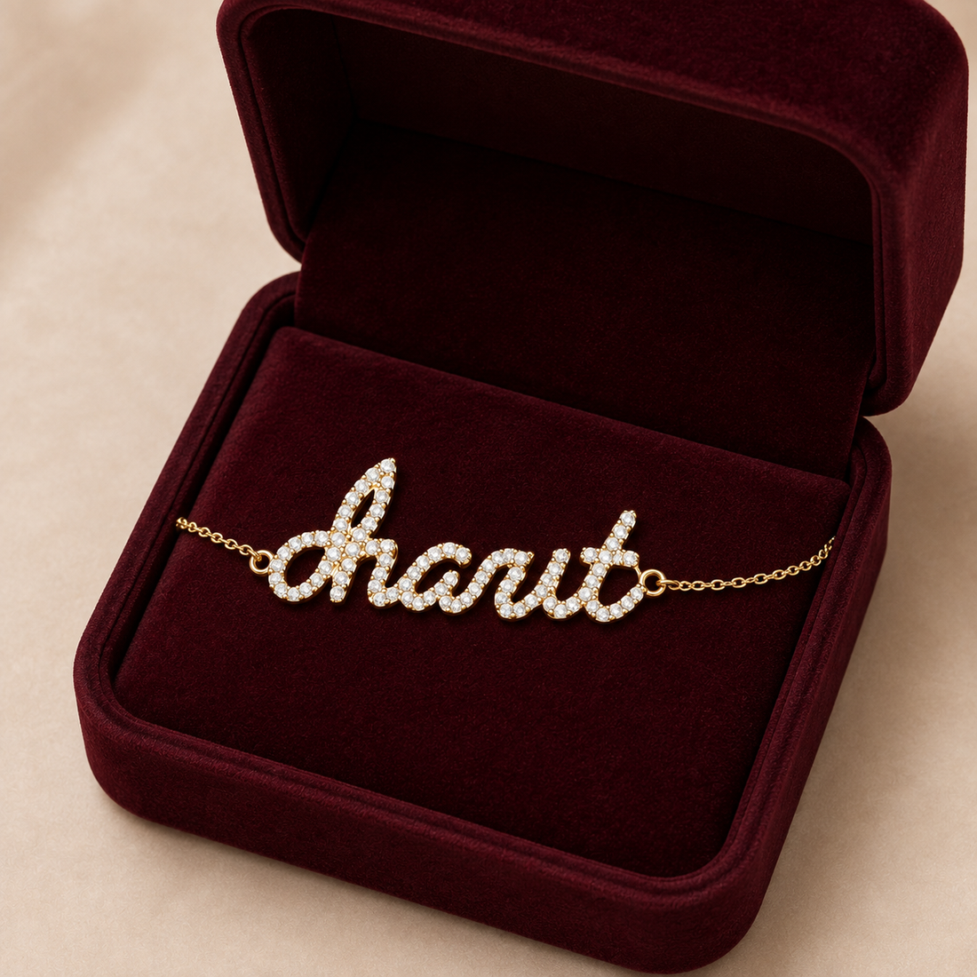 A personalized name bracelet spelling charit  in script, set with clear stones on a gold chain, displayed inside an open burgundy velvet jewelry box on a beige background.
