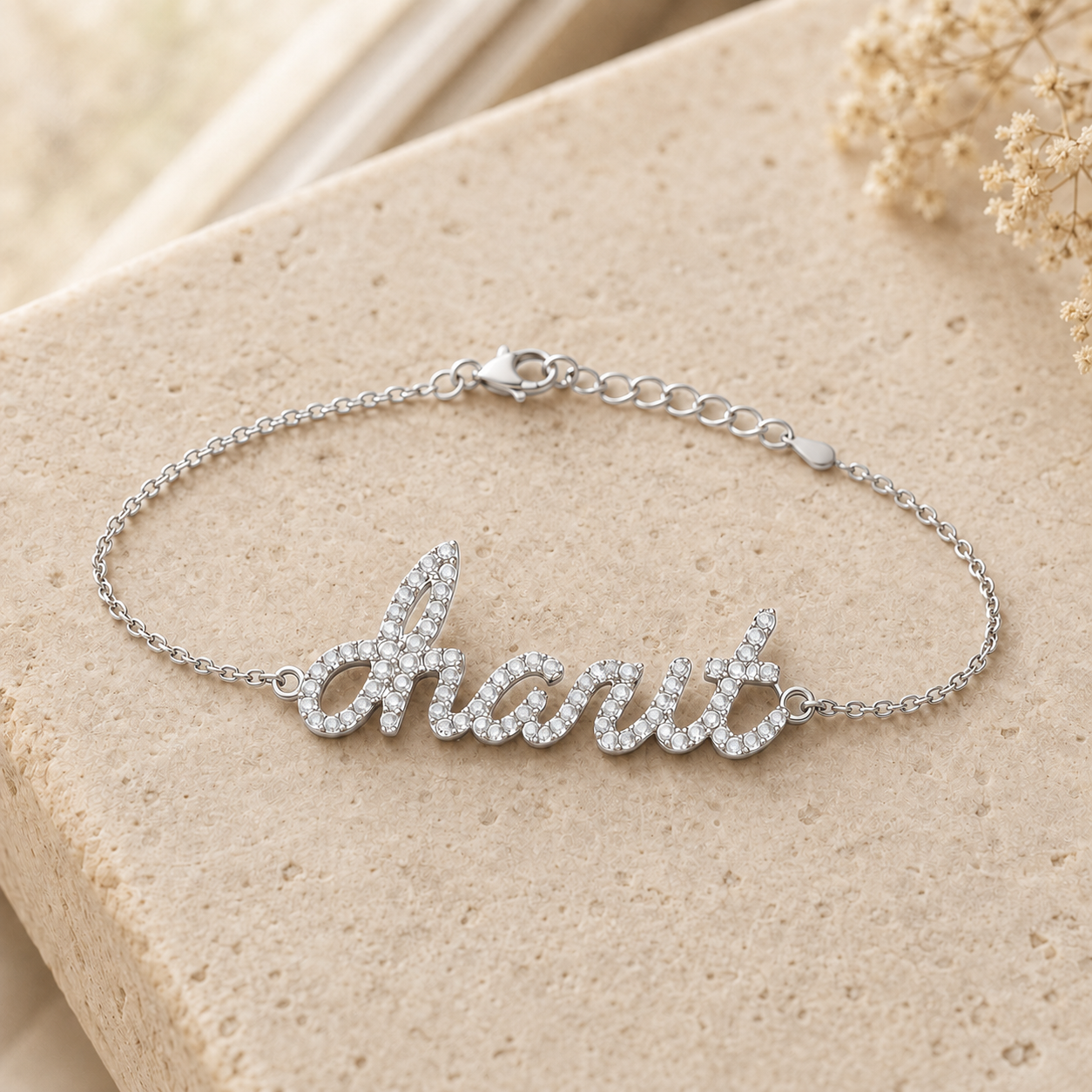 A silver script nameplate bracelet with clear rhinestones spelling charit resting on a textured beige stone surface with dried flowers in the background.