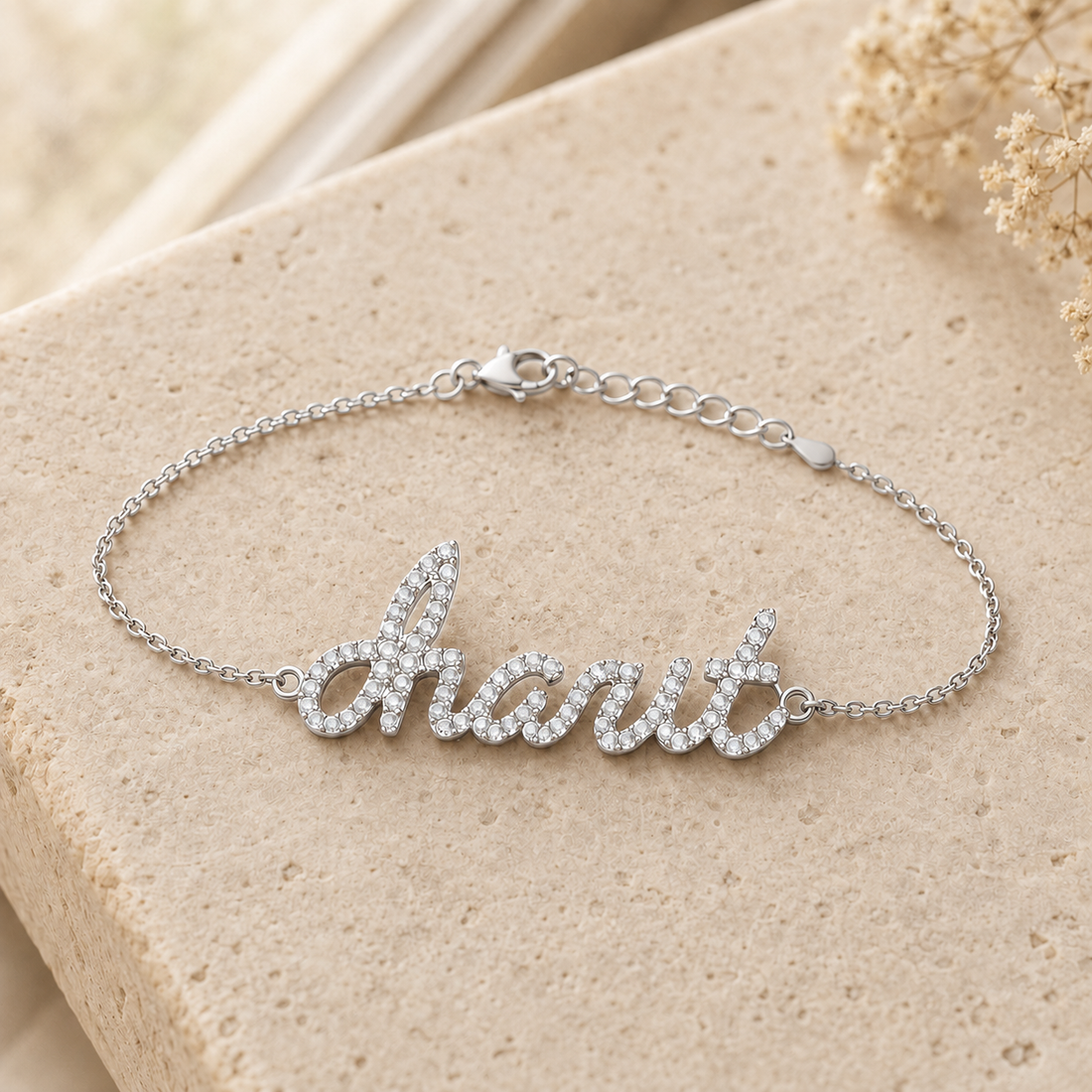 A silver script nameplate bracelet with clear rhinestones spelling charit resting on a textured beige stone surface with dried flowers in the background.