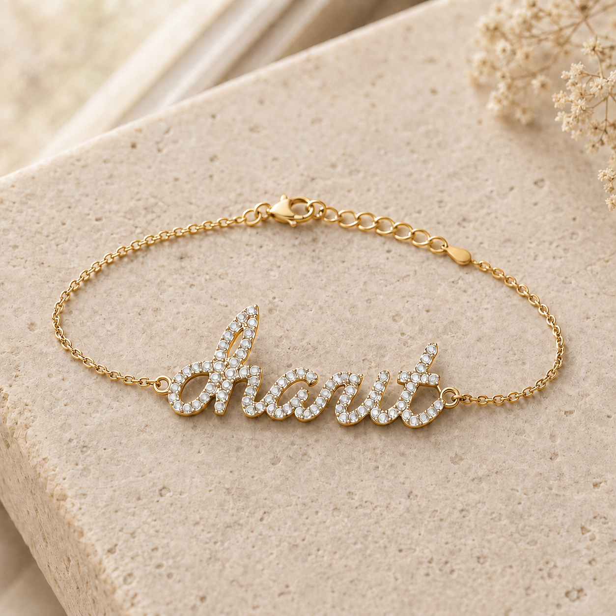 A gold chain bracelet with a cursive nameplate spelling charit in clear rhinestones, displayed on a textured beige stone surface.