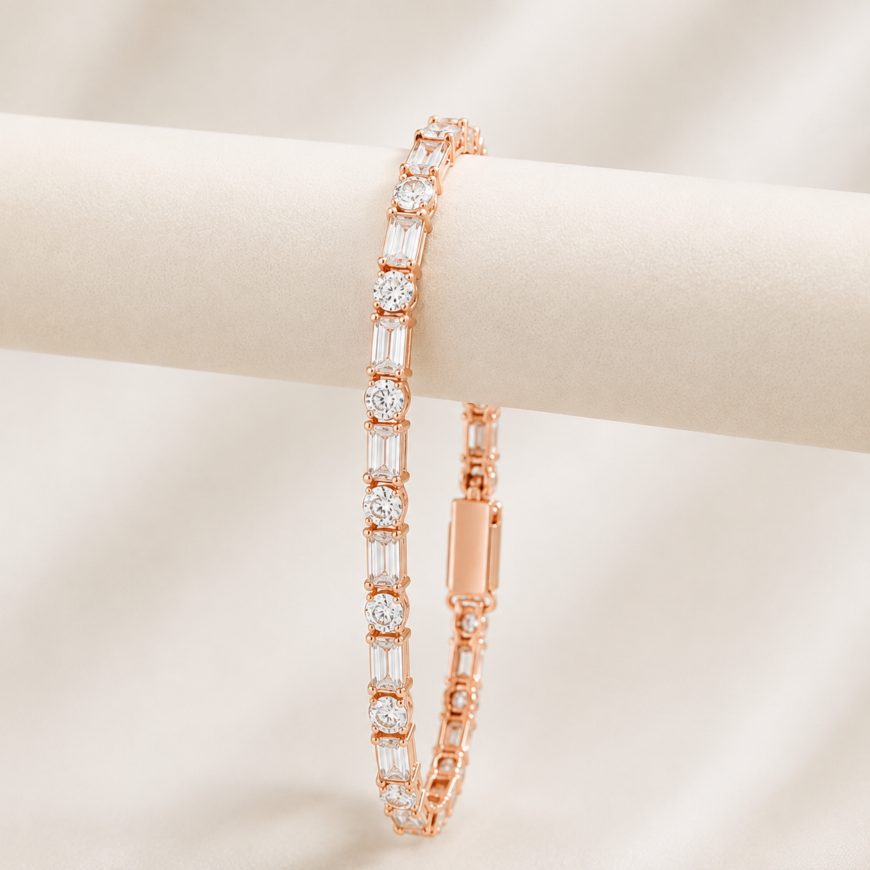 A rose gold tennis bracelet with round and baguette-cut clear stones, displayed on a cream-colored cylindrical jewelry stand against a soft beige background.