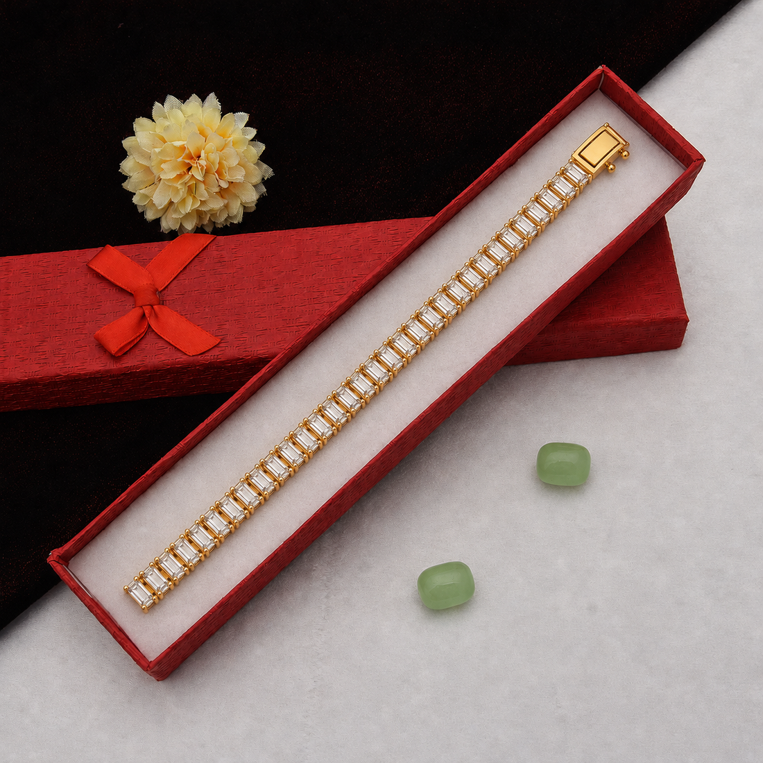 A gold-toned bracelet with rectangular clear stones arranged in a row, presented inside an open red jewelry box. A red gift box with a bow and a pale yellow flower sit behind it on a light surface, with two small green stones nearby.