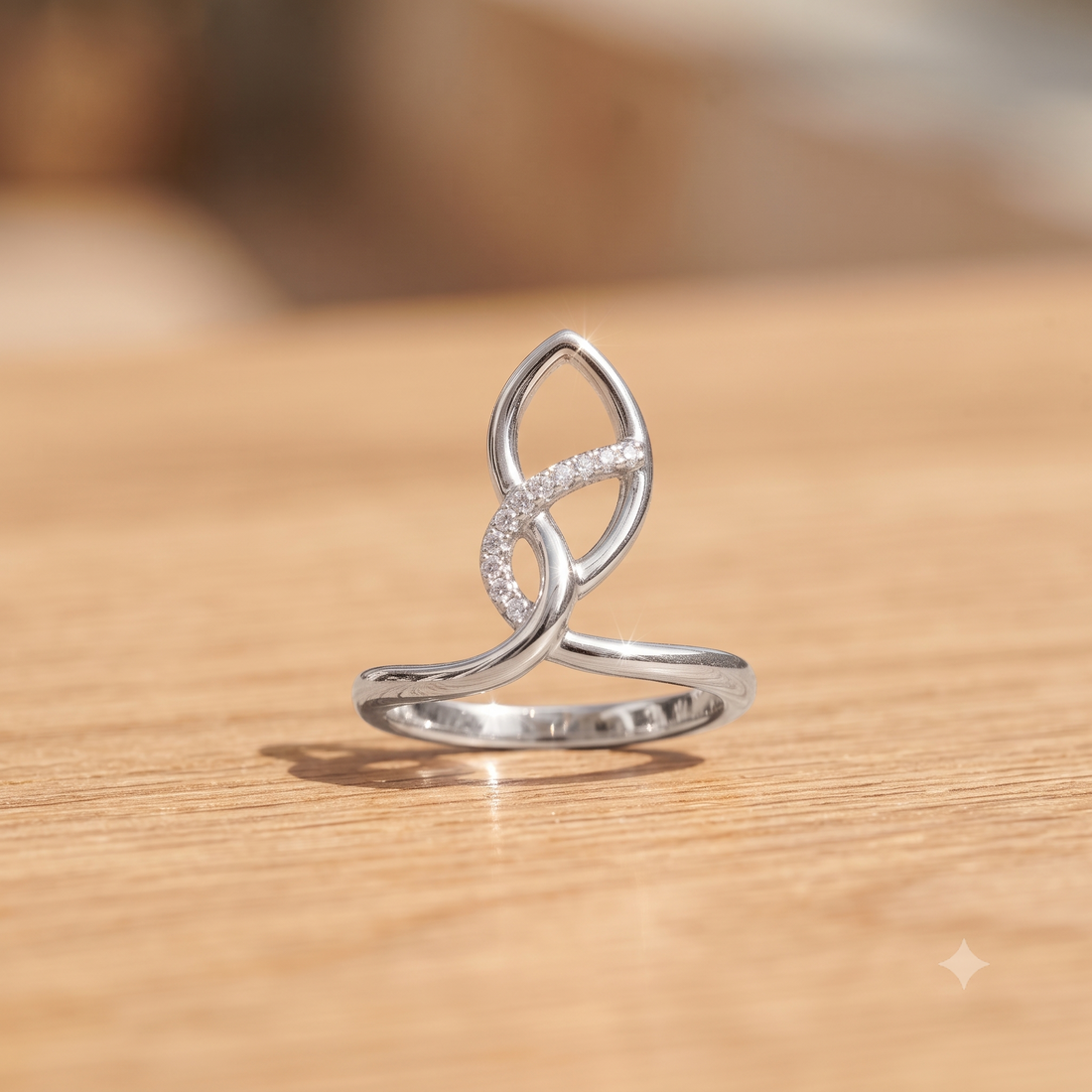 A silver open-band ring with a stylized infinity symbol set with small clear stones, resting on a light wood surface.