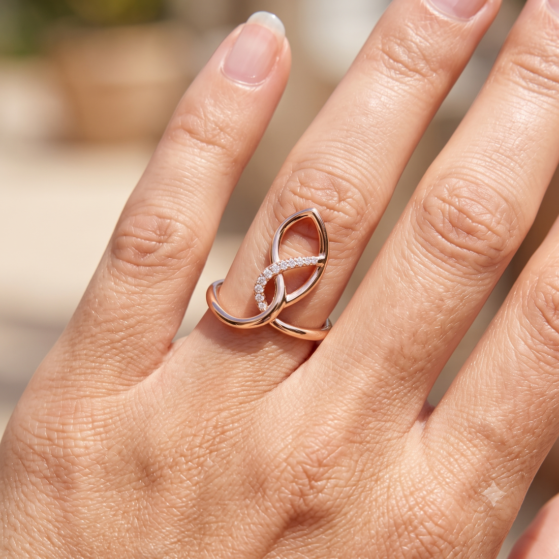 A close-up of a hand wearing a rose gold infinity-style ring with a row of small clear stones across the center, set against a softly blurred outdoor background.