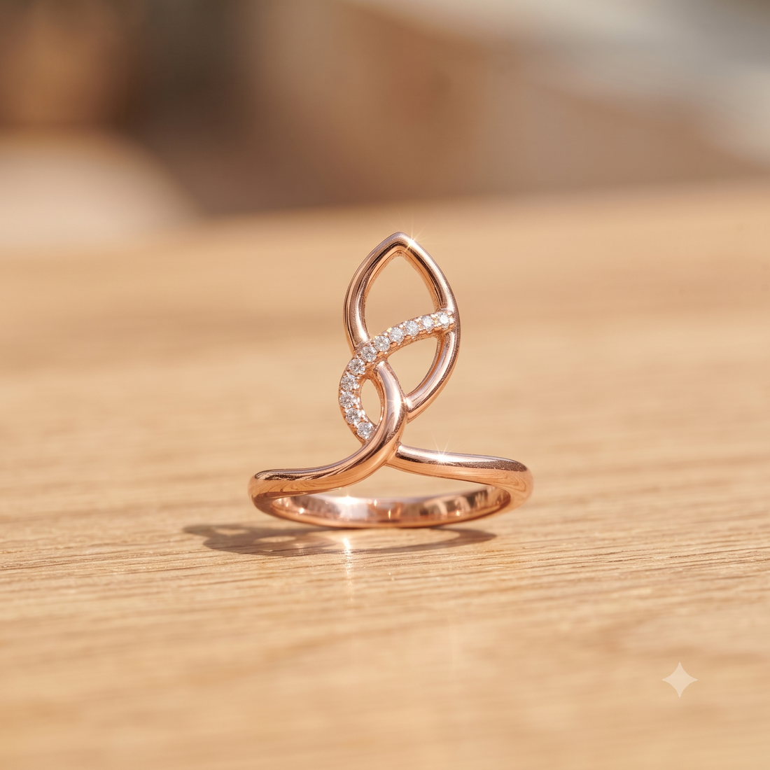 A rose gold ring with a stylized knot design and a curved section paved with small clear stones, resting on a light wood surface.
