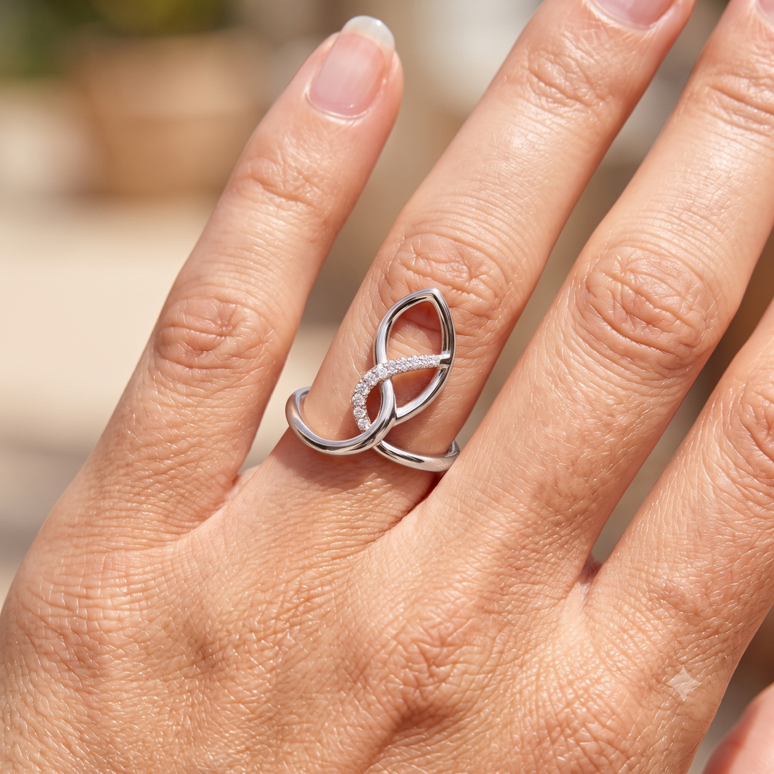 A close-up of a hand wearing a silver ring with two interlocking loop shapes, one of which is set with small clear stones, against a softly blurred outdoor background.