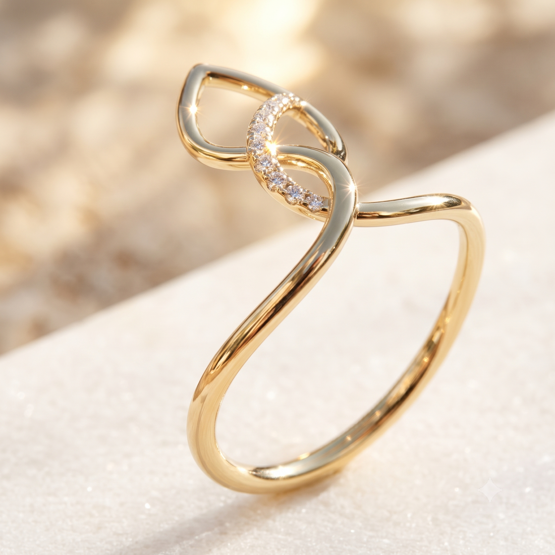 A gold ring with a stylized bow-like top featuring a curved row of small clear stones, photographed on a soft light beige surface with a blurred warm background.