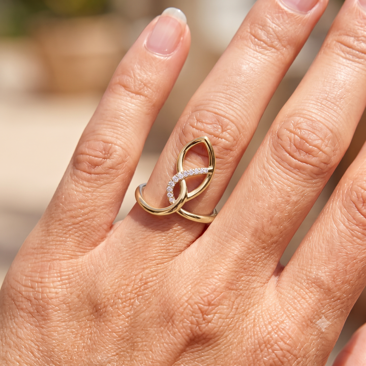 A close-up of a hand wearing a gold ring with two interlocking curved bands, one of which is set with small clear stones, photographed in bright natural light.