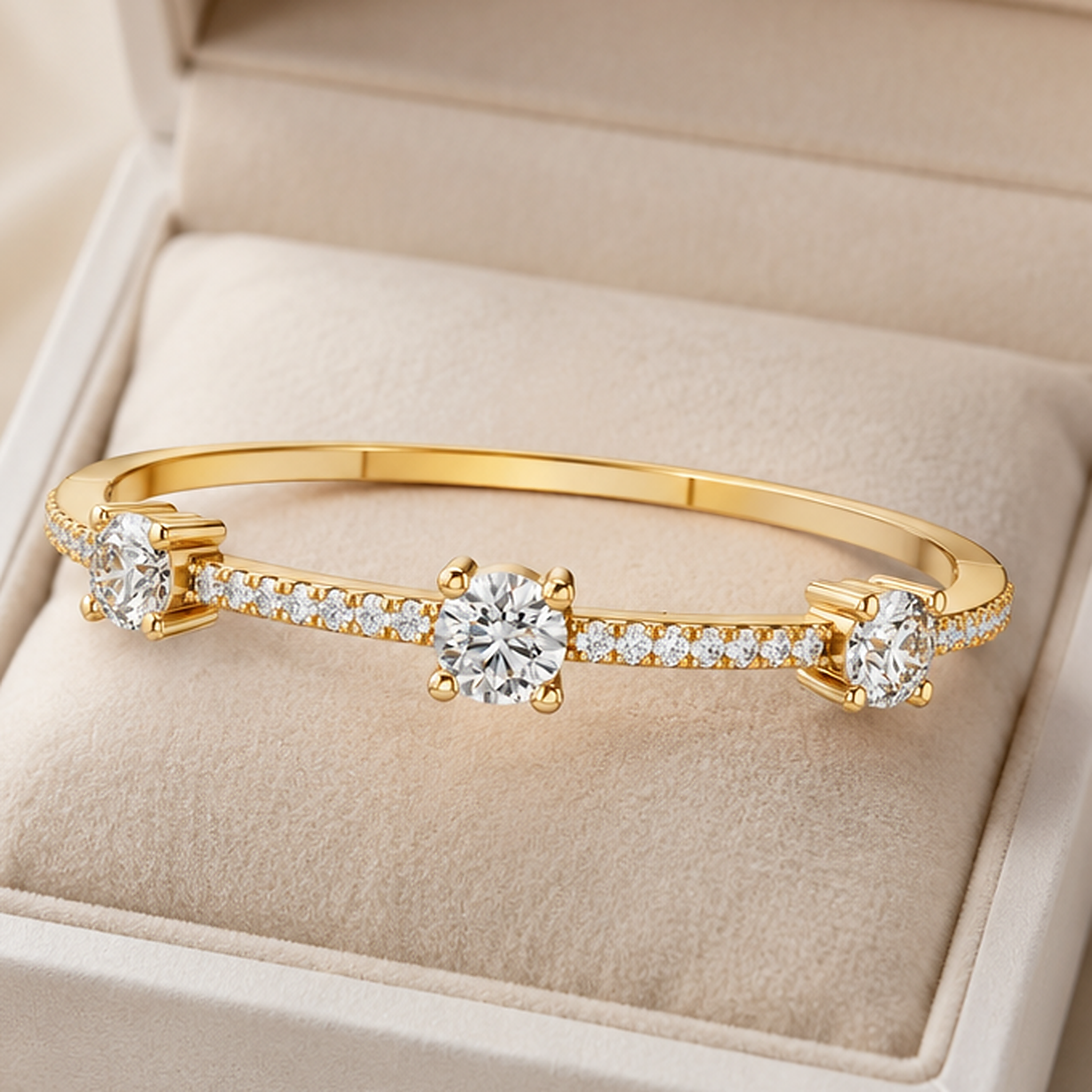 A gold bangle bracelet with three round-cut clear center stones and small pavé accents, displayed inside a beige jewelry box.