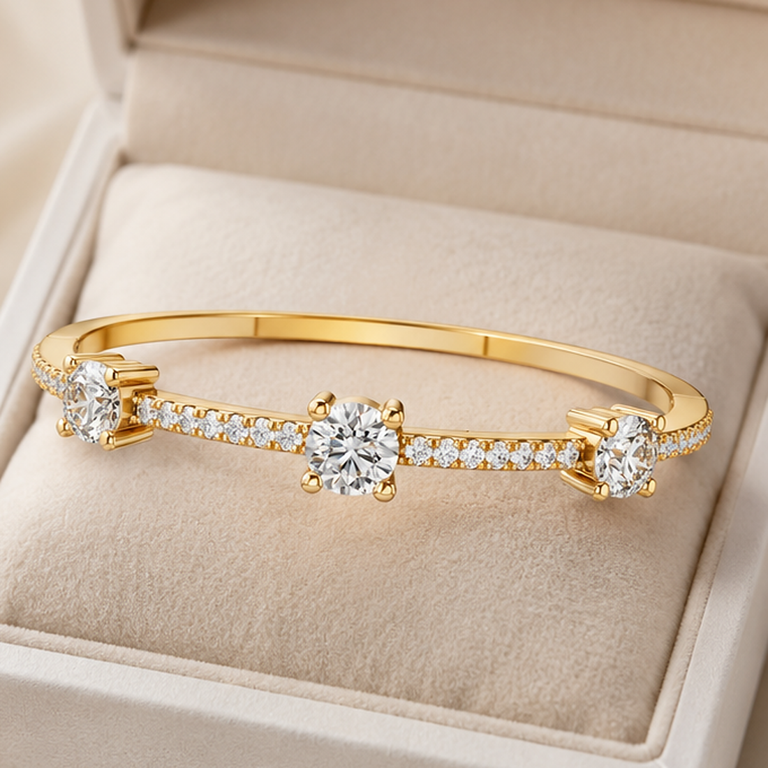 A gold bangle bracelet with three round-cut clear center stones and small pavé accents, displayed inside a beige jewelry box.