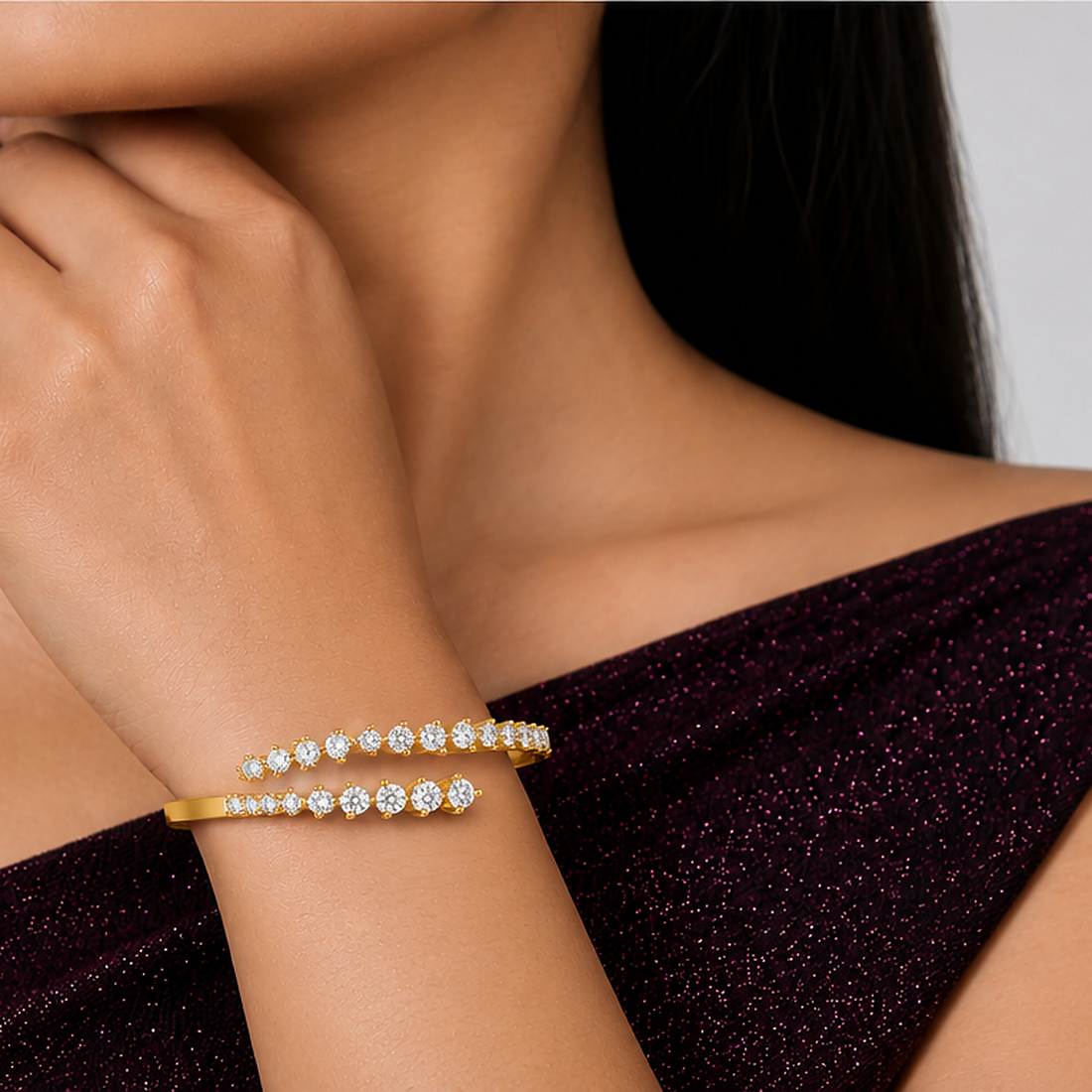 A close-up lifestyle shot of a model wearing a gold bangle bracelet with rows of sparkling clear gemstones. The model is wearing a dark plum off-the-shoulder top, and the background is a clean, neutral gray.